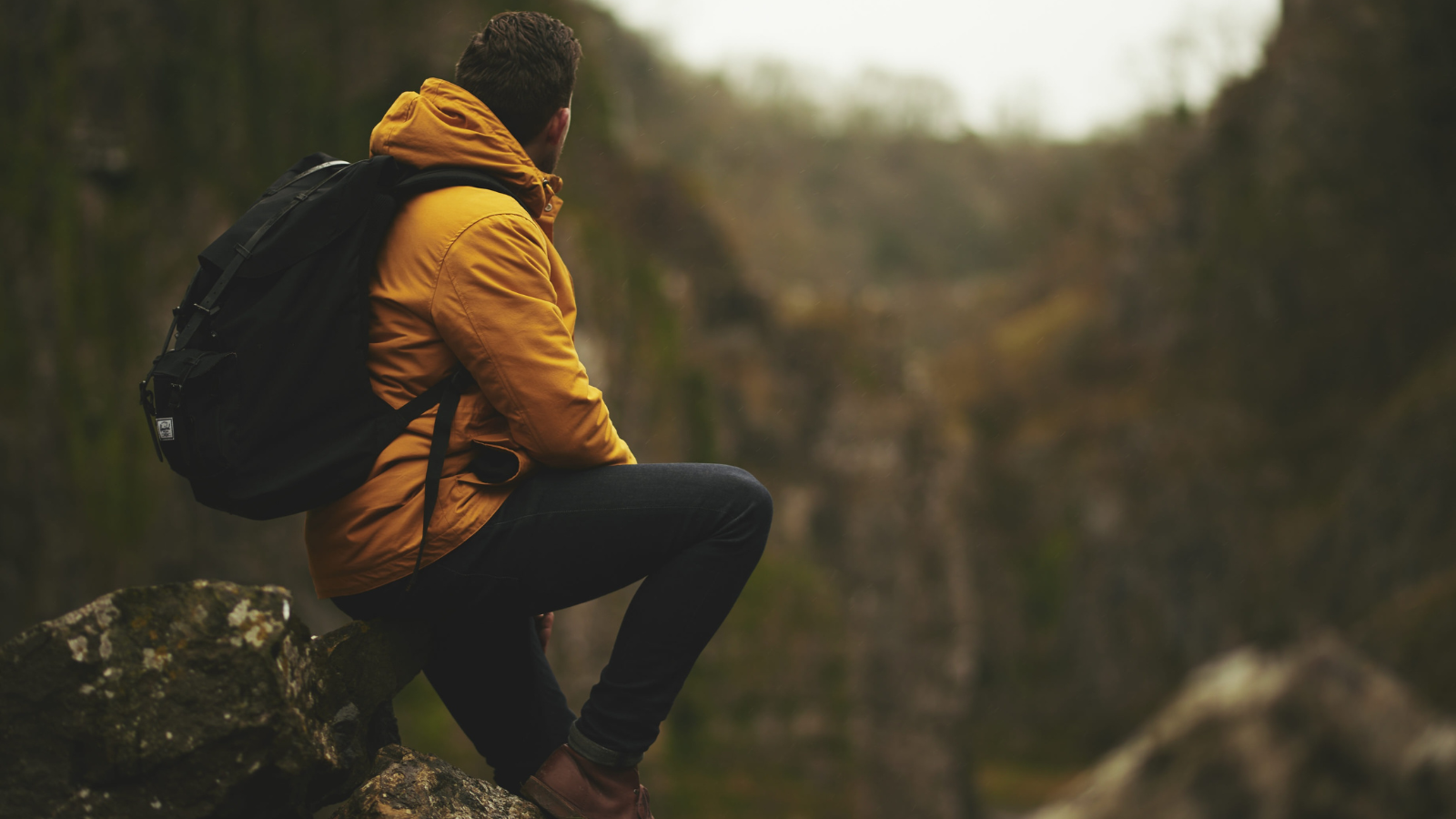 A hiker sits on a rock in a rugged mountain landscape, wearing a yellow jacket and looking out over the hills